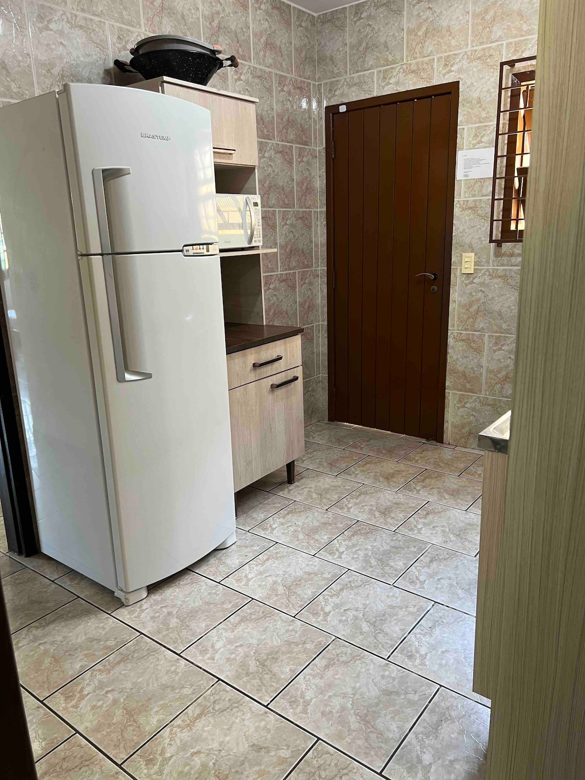 A functional kitchen space is depicted, featuring light wooden cabinetry and a large white refrigerator. The tiled floor is complemented by patterned walls, enhancing the cozy environment. A microwave and a small countertop provide additional cooking convenience.