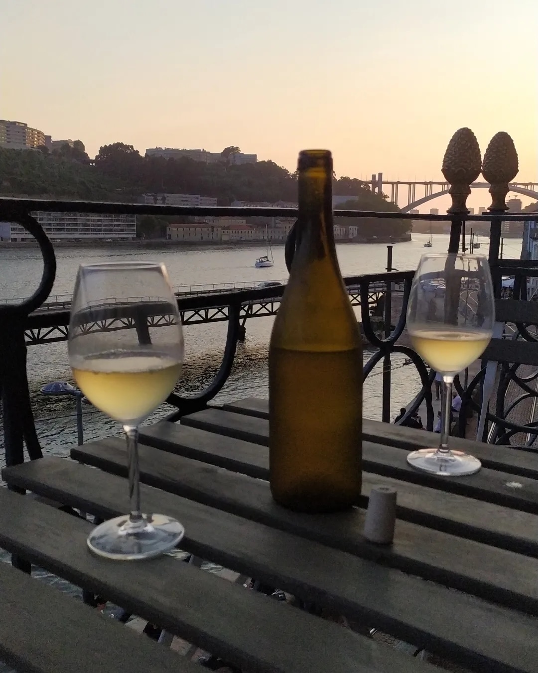A table is set with two glasses of white wine, paired with a partially filled brown bottle, overlooking the river. A scenic waterfront is visible, highlighting the serene atmosphere at sunset. The intricate railing adds a touch of elegance to the balcony view.
