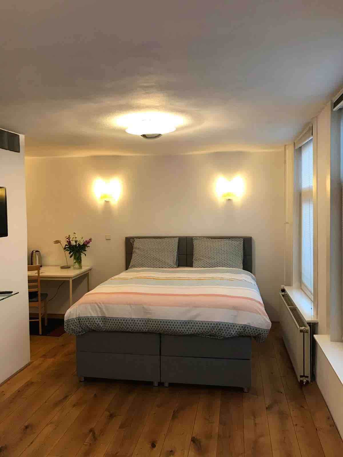 A spacious room is brightened by soft lighting, featuring a double bed with a patterned duvet. A small table is positioned nearby, accompanied by a kettle. Wooden floors enhance the room's warmth, while large windows allow natural light to fill the space.