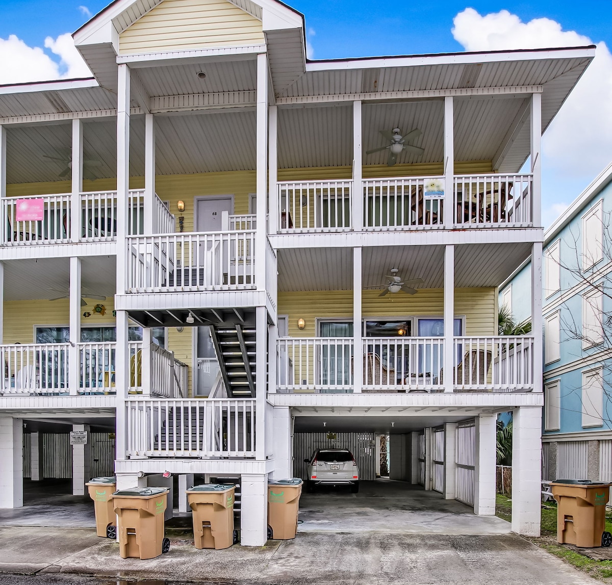 The exterior of the cottage features a three-level structure with a light yellow facade and white railings. Two covered parking spaces are visible under the building, along with outdoor trash receptacles. A staircase on one side leads to the upper levels, and balconies are shown.