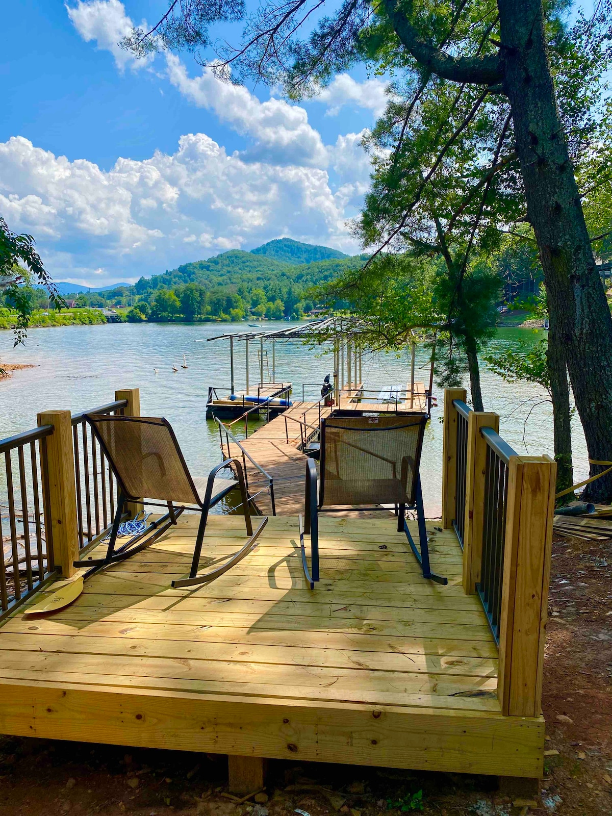A wooden deck extends over the water, featuring two chairs positioned for relaxation. The boat dock is visible, with boats moored nearby. Lush trees and rolling hills frame the serene lake under a partly cloudy sky.