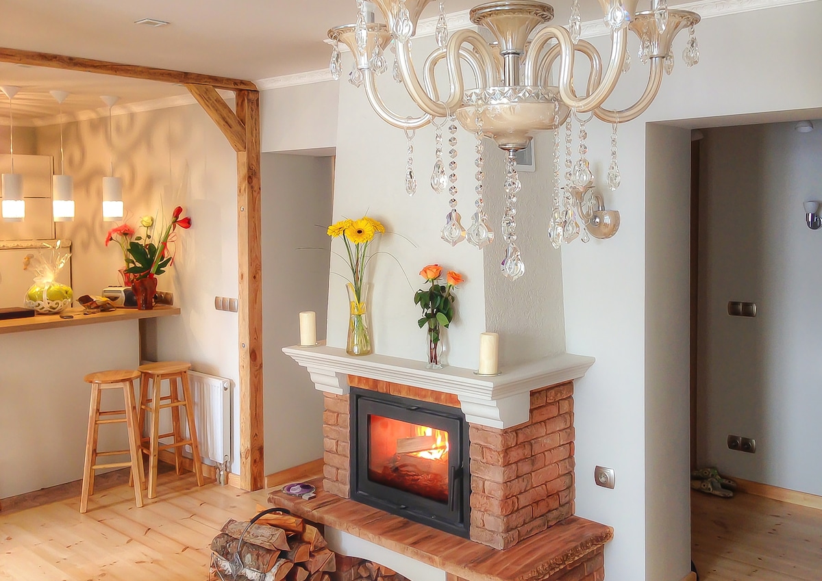 An elegantly designed living area features a cozy fireplace surrounded by a stone mantel. A chandelier with hanging crystals adds a touch of sophistication. Bright flowers on a bar counter and warm wooden flooring enhance the inviting ambiance of the space.
