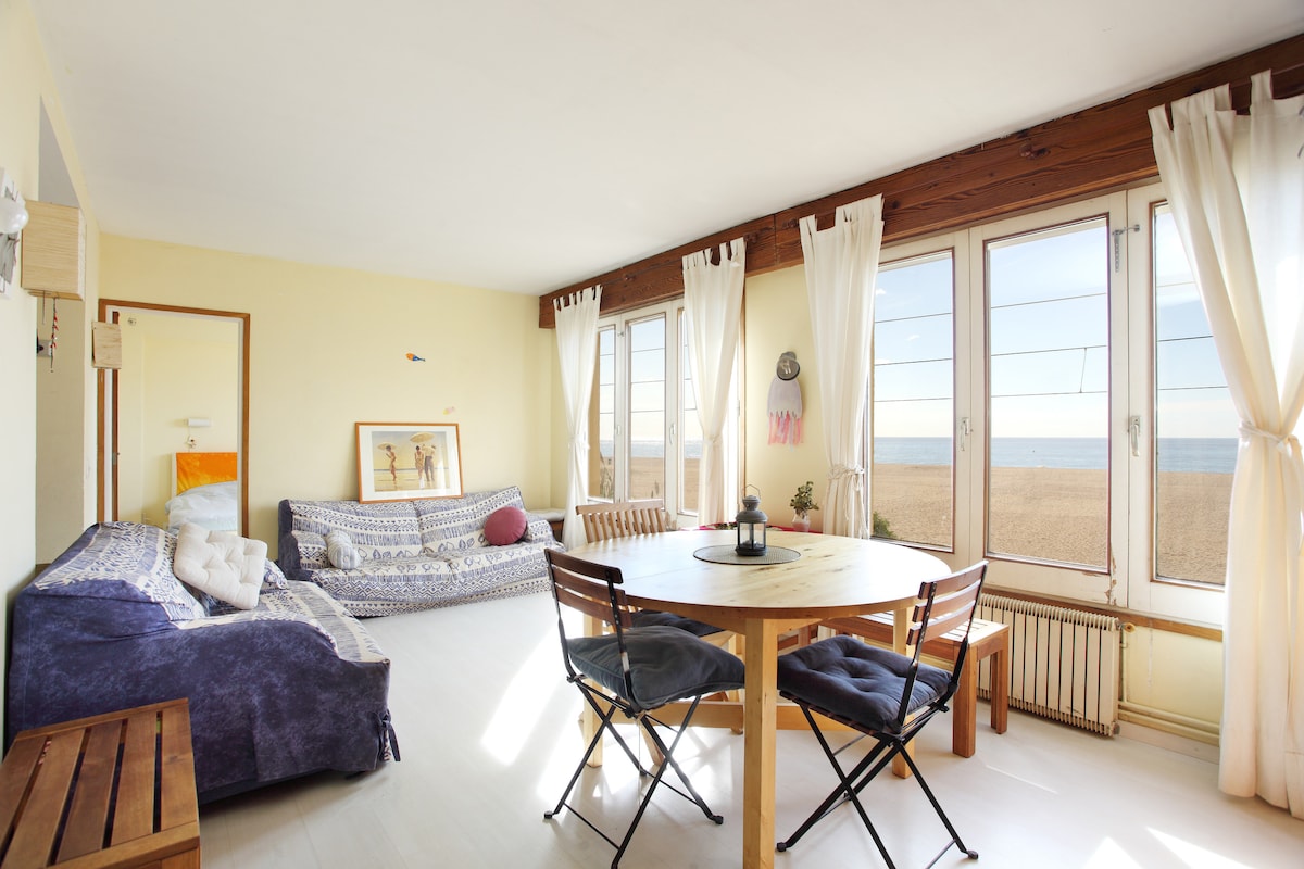 Natural light fills the spacious living area, featuring large windows that provide beach views. A dining table is set for four, accompanied by cozy seating. Soft textiles add a calming touch to the room, while light curtains gently sway with the breeze.