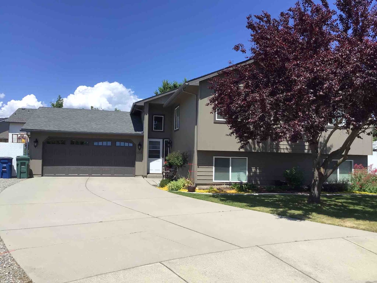 The exterior features a two-story tan house set on a spacious driveway, complemented by neatly landscaped flower beds. A striking red tree adds a touch of color to the front yard under a clear blue sky.