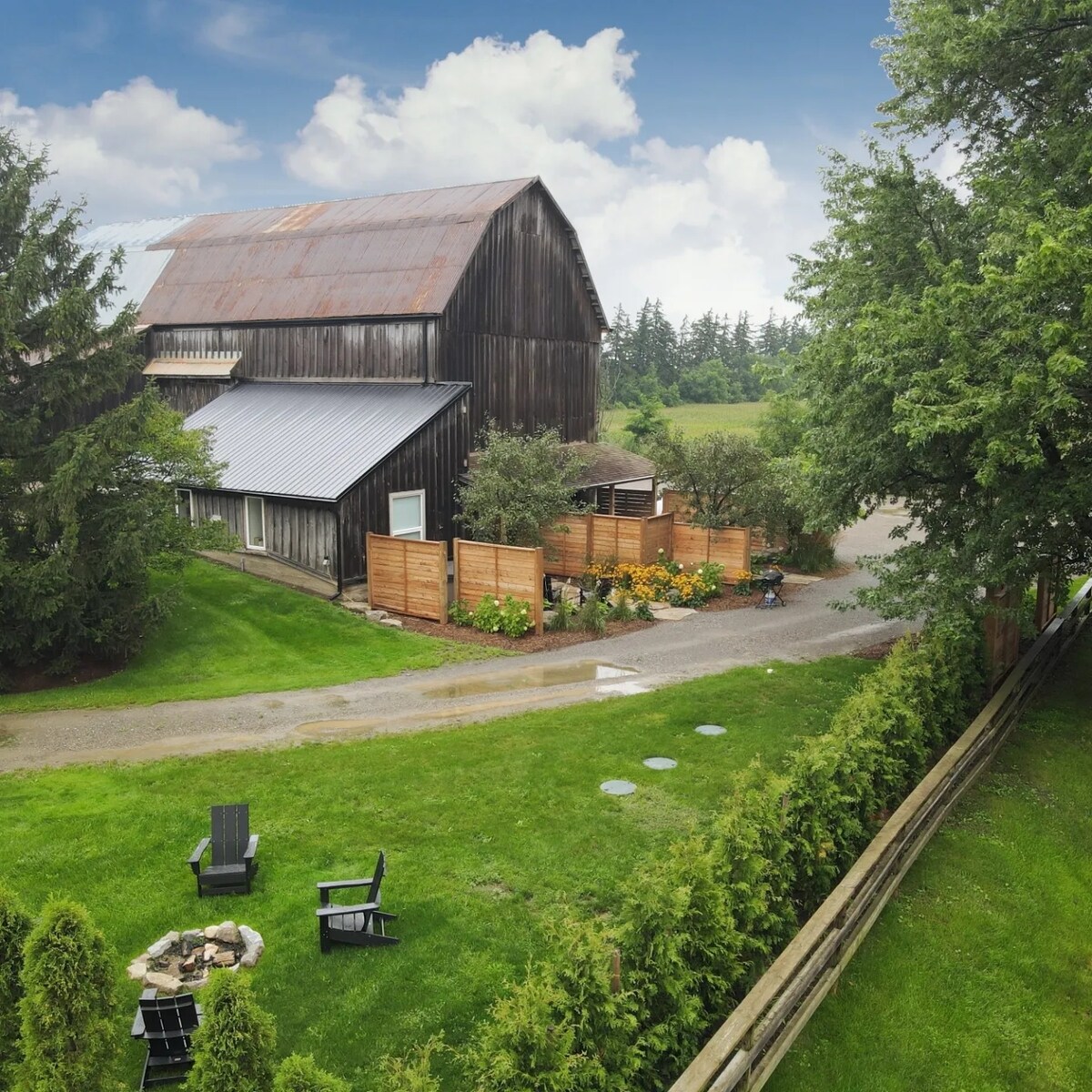 A rustic barn structure with a weathered roof is set against a backdrop of greenery. A pathway leads to the barn, bordered by wooden fencing and plant beds. A fire pit surrounded by Adirondack chairs is visible on the lawn, inviting relaxation in the outdoor space.