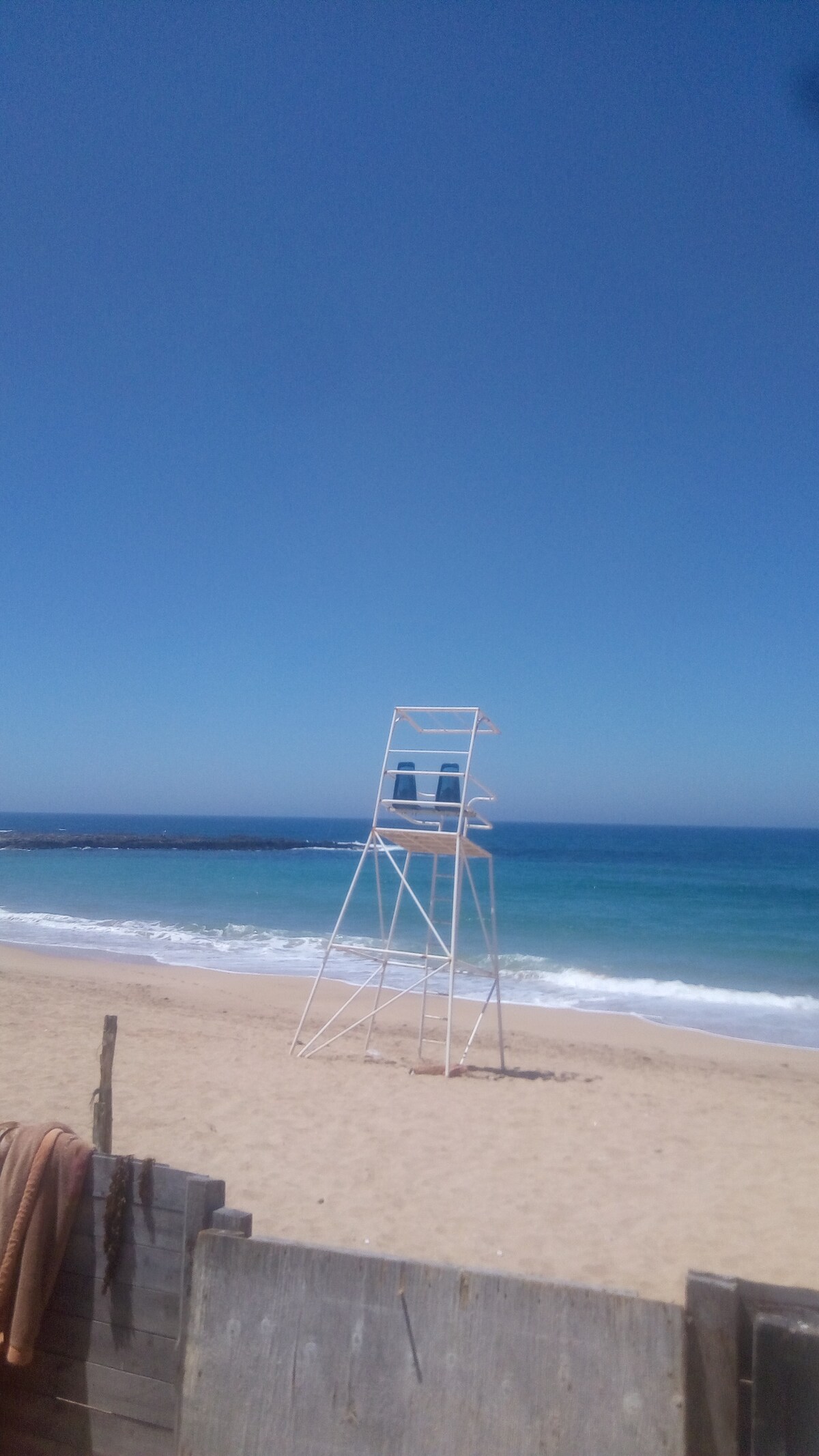 A lifeguard tower is positioned on a sandy beach, overlooking gentle waves lapping at the shore. A clear blue sky stretches above, complementing the serene ocean hues. Wooden structures are partially visible in the foreground, hinting at the coastal environment.