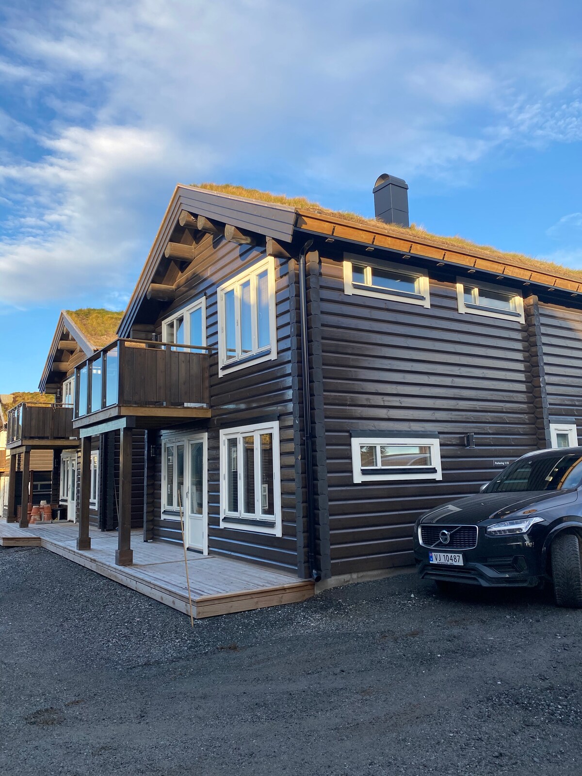 The exterior of the duplex cabin is showcased, featuring a dark wood facade with a grass-covered roof. A spacious balcony is visible on the upper level, and a driveway accommodates a parked vehicle. The surrounding area is gravel, with clear blue skies above.
