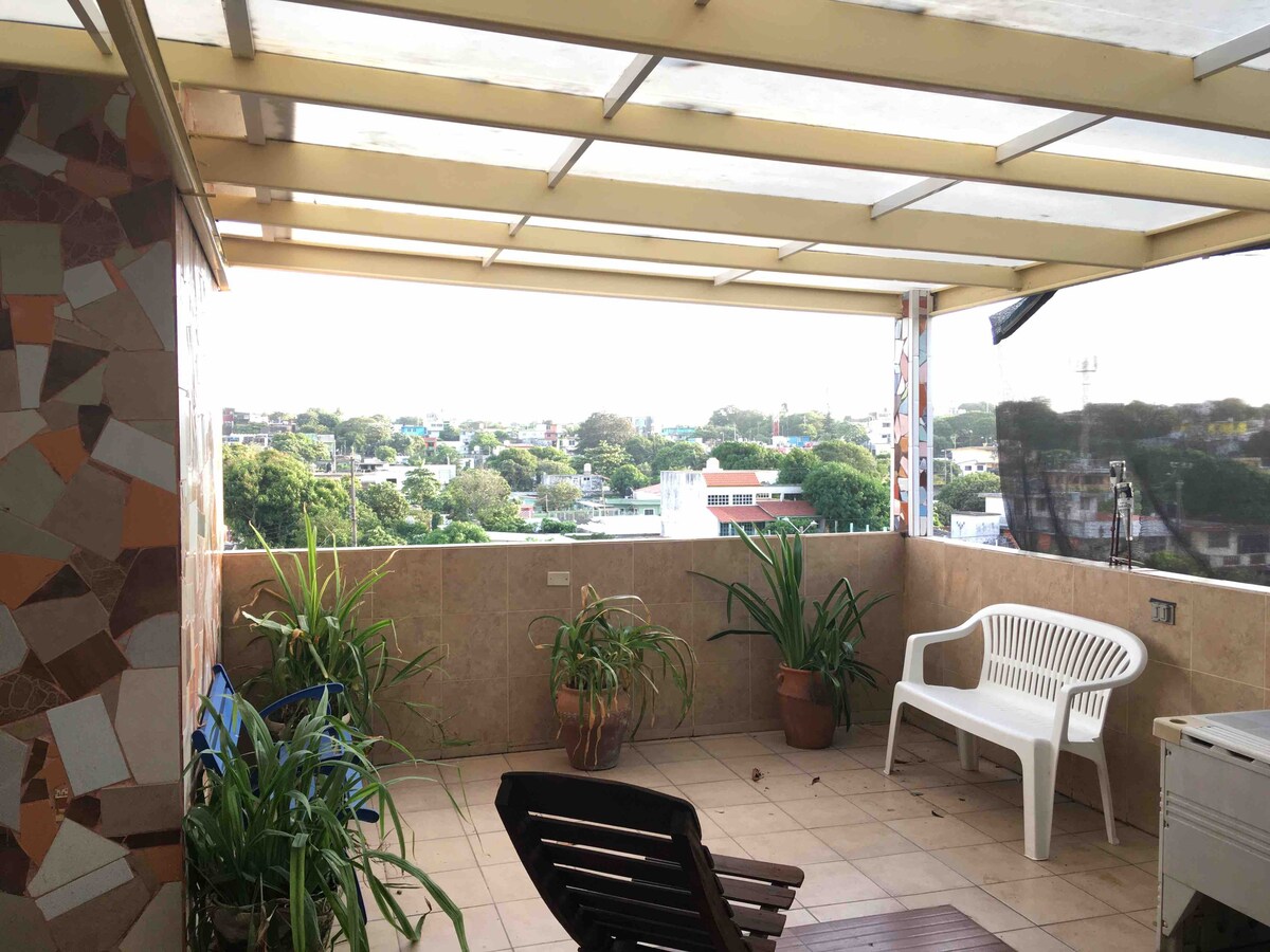 A spacious terrace is shown, featuring a partial roof and offering a view of the surrounding cityscape. Potted plants are placed along the tiles, while a white chair and a wooden lounge chair provide seating options. The area is well-lit with natural light.