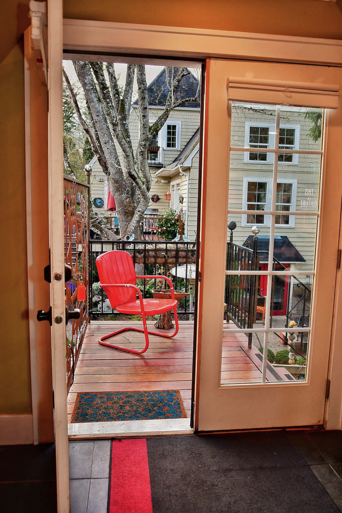 A view from the studio's french doors opens to a small outdoor sitting area featuring a red chair on a wooden deck. Green trees are visible, along with nearby buildings showcasing varied architectural styles.