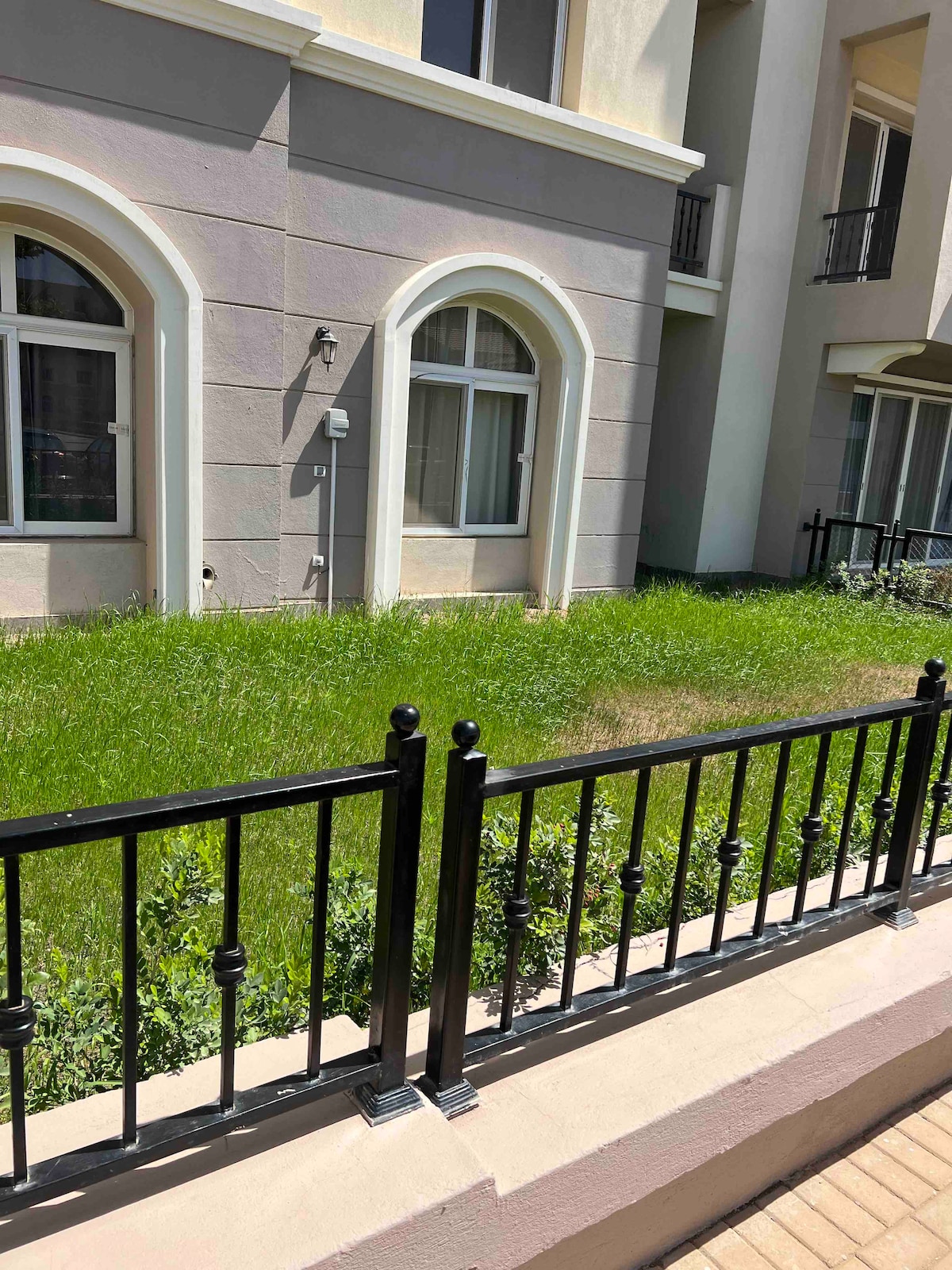 A well-maintained green lawn is bordered by a black wrought iron fence, providing a defined outdoor area. Large arched windows of the adjacent building create a visual rhythm along the exterior, allowing natural light to enter the space.