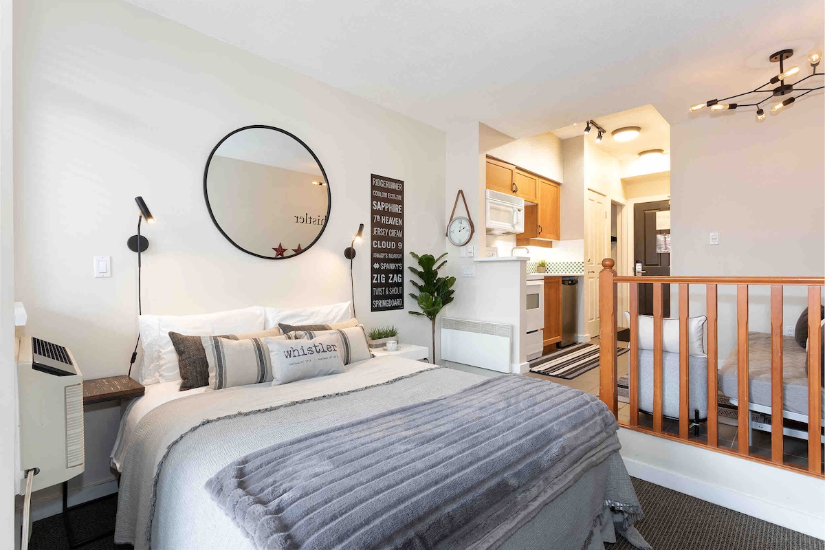 A cozy studio space features a Queen bed adorned with neutral linens and a plush throw blanket. A round mirror hangs above the bed. The kitchenette is visible in the background, equipped with essential appliances. A seating area is present to the right, offering additional comfort.