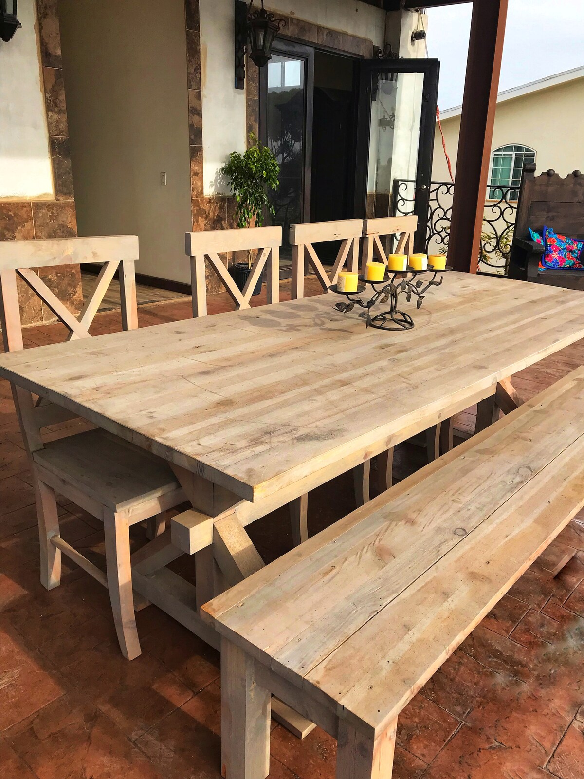 A wooden dining table with a rustic finish is surrounded by matching chairs and a bench. Six yellow candles are arranged on an ornate candle holder at the center. The outdoor space features a backdrop of stone walls and large glass doors leading to the interior.