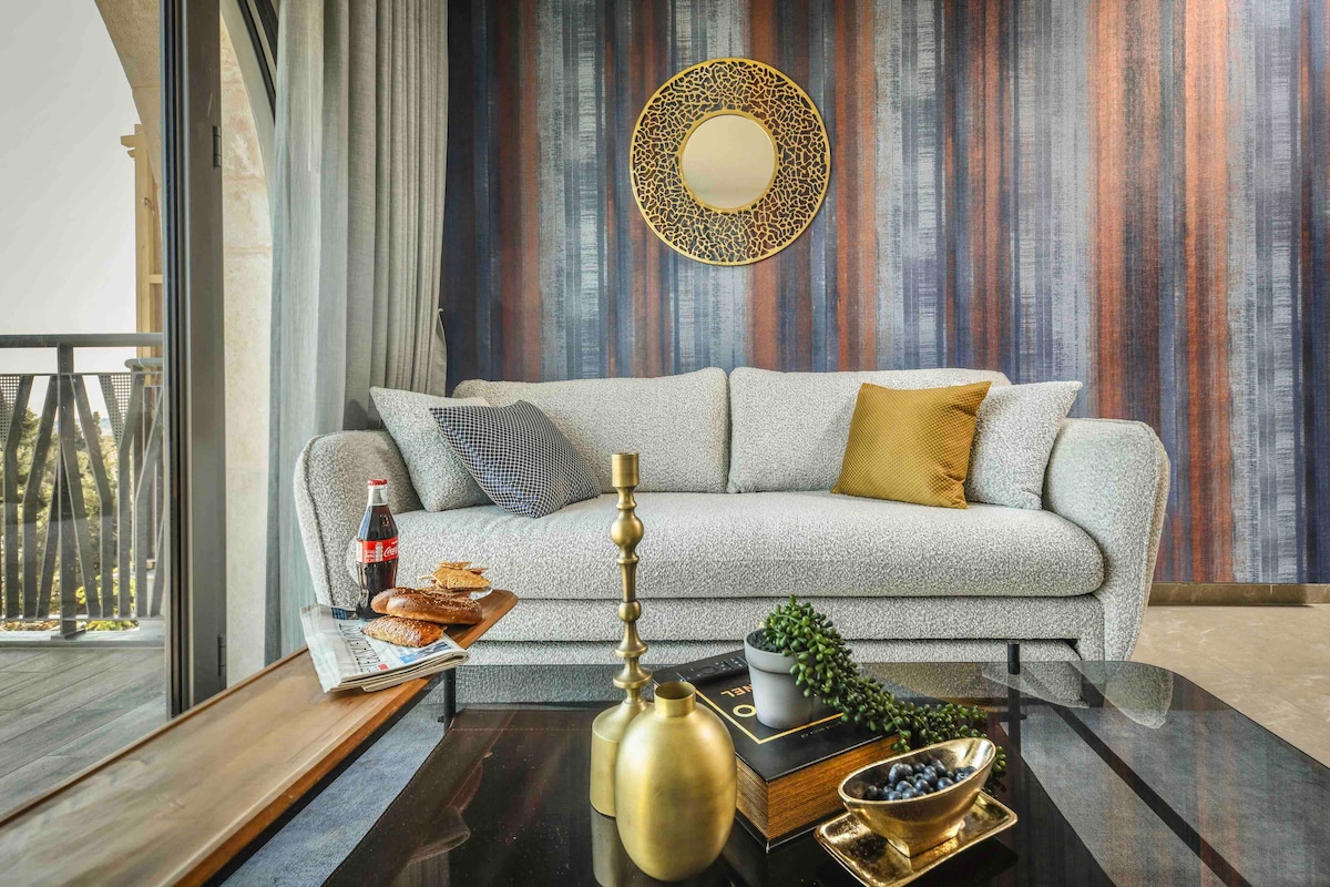 A light-colored, textured sofa is positioned in front of a glass coffee table, showcasing decorative items. A round mirror hangs above the couch, reflecting natural light. The striped wall in varying shades adds depth to the airy living space, which features large windows with views of the balcony.