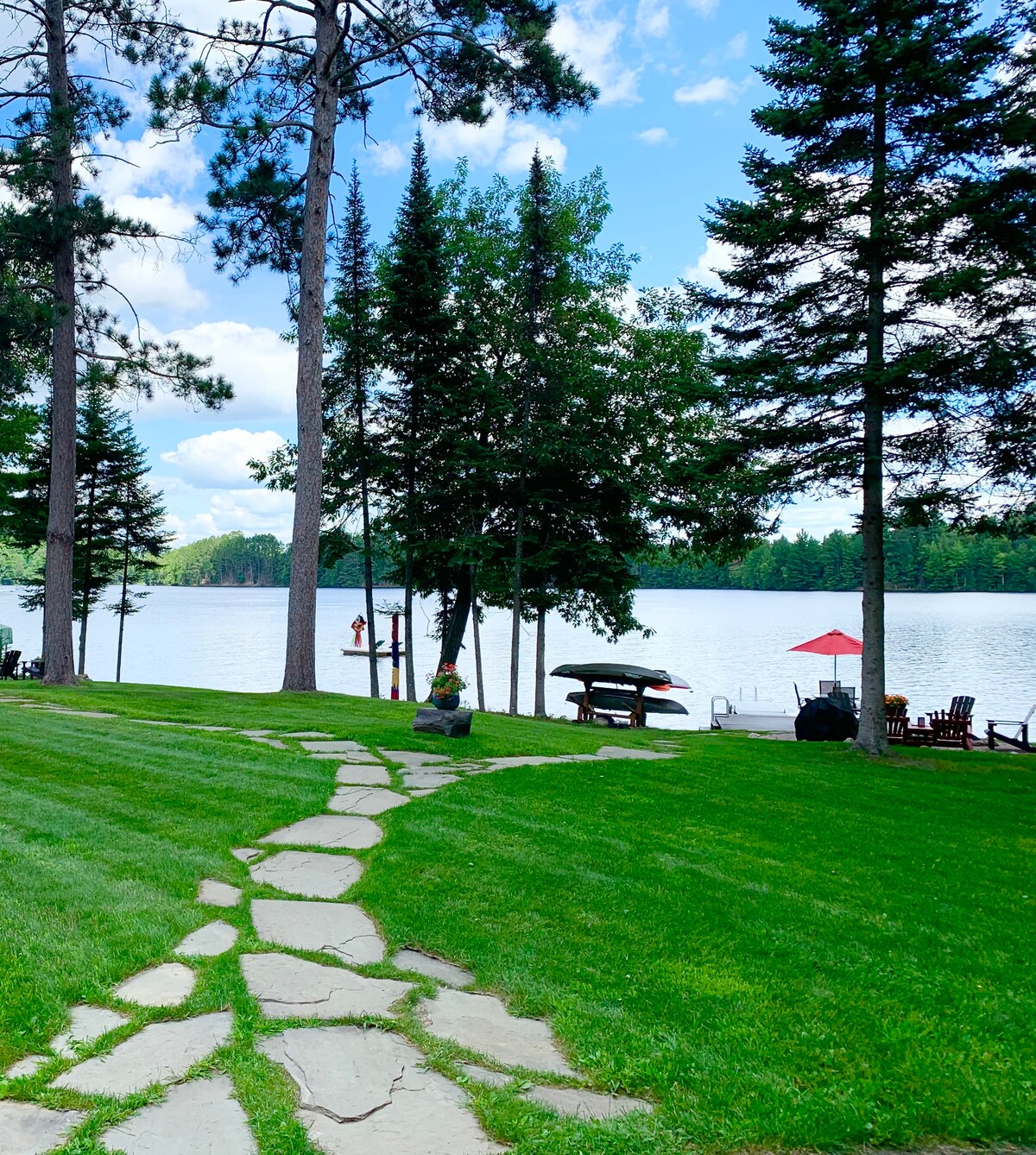 A stone pathway leads through lush green grass to a tranquil lake, surrounded by tall pine trees. A wooden pier extends into the water, with a red umbrella over a seating area nearby. Guests can be seen enjoying the lake in the distance.
