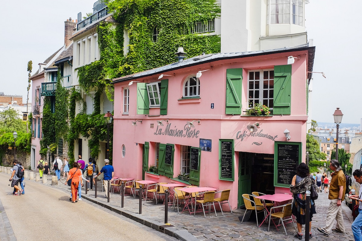A charming café, La Maison Rose, is situated on a cobblestone street adorned with vibrant pink walls and green shutters. Small outdoor tables and chairs create an inviting space for guests. Lush greenery climbs the side of the building, adding to the serene Parisian ambiance.