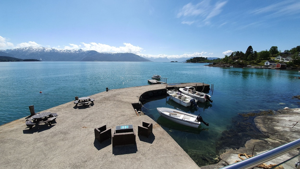 A concrete pier extends into the tranquil waters of the Hardangerfjord, featuring several boats docked alongside. Outdoor seating areas with dark wicker furniture are visible, overlooking the scenic view of mountains and clear blue sky.