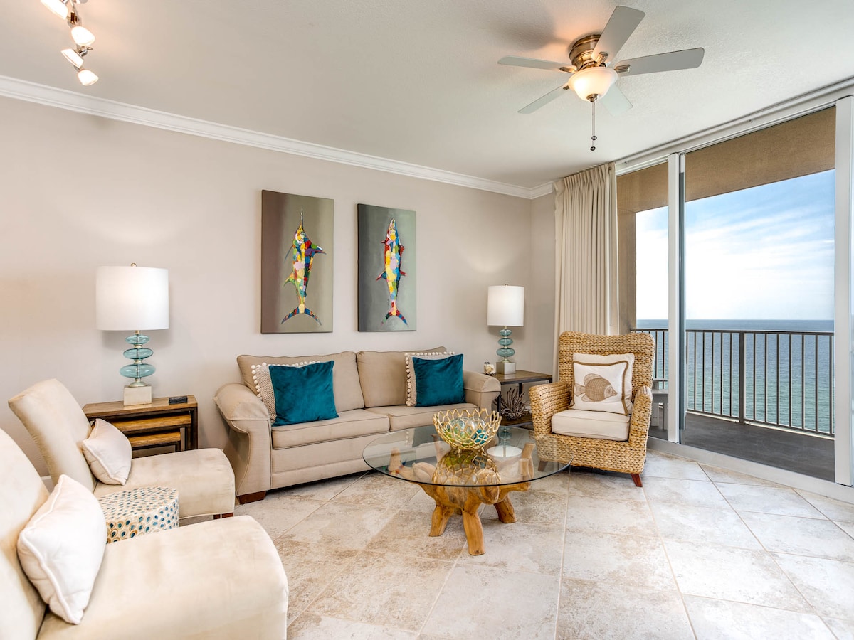 A bright living area is showcased with large windows offering views of the Gulf. A comfortable sofa and chairs are arranged around a glass coffee table atop tiled flooring. Colorful artwork adorns the walls, with natural light spilling in through the sliding doors.