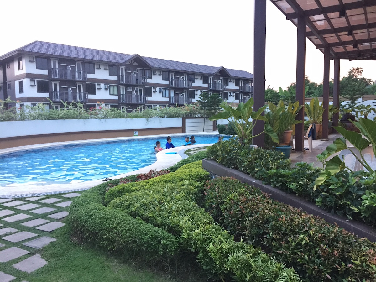 A swimming pool surrounded by lush greenery and well-maintained landscaping is visible. Guests are seen enjoying the pool, while modern condominium buildings provide a backdrop. The area features a covered seating space, enhancing the outdoor experience.