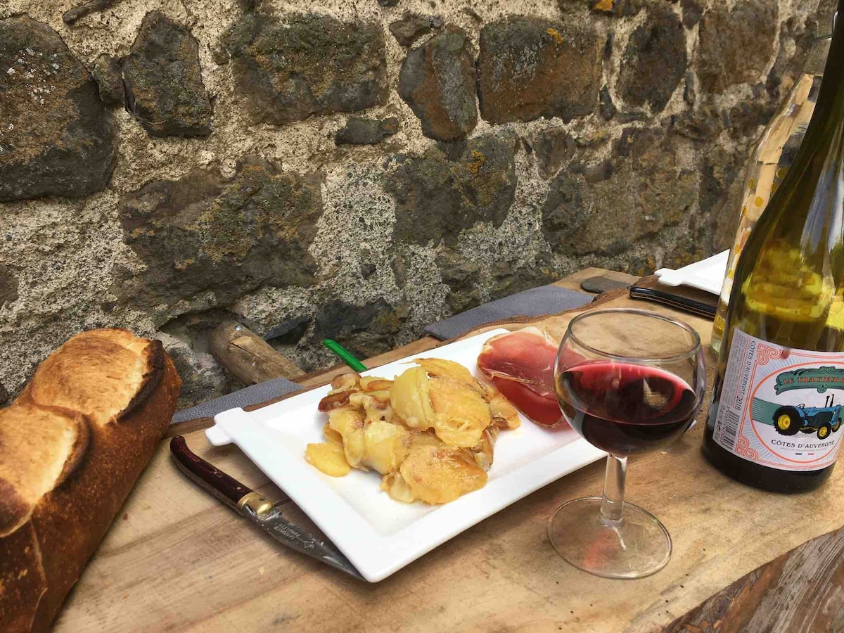 A wooden table displays a plate of golden sautéed potatoes alongside sliced meat and a glass of red wine. A bottle of white wine sits nearby, with a rustic stone wall in the background, adding texture to the setting.