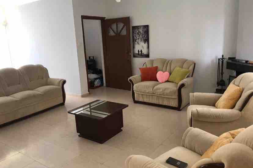 A bright living area features two cream sofas and two armchairs arranged around a low coffee table. A warm atmosphere is provided by light-colored walls and soft seating. A door leads to another room, while a wall adorns a serene piece of artwork.
