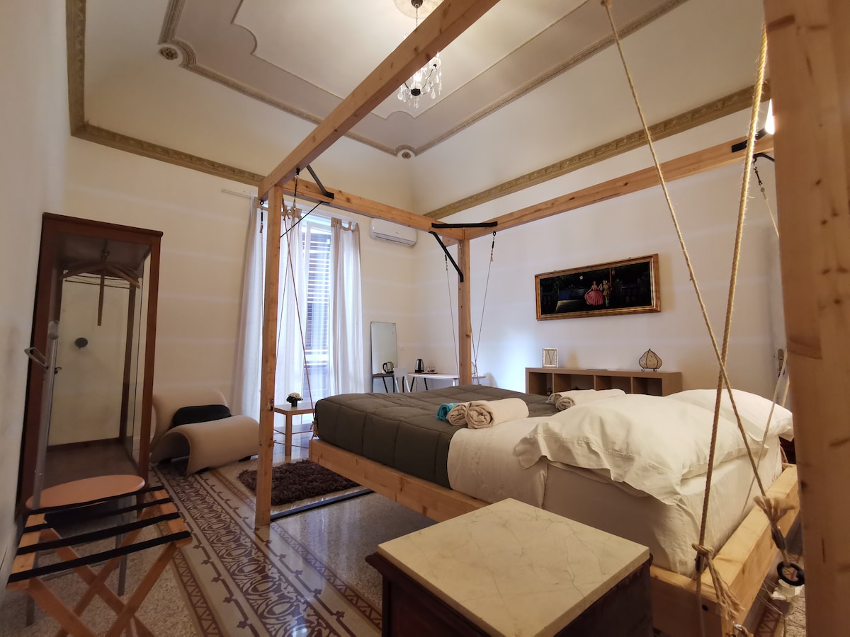 A spacious room featuring a suspended bed, crafted with wooden beams. Natural light filters through sheer curtains, illuminating the elegantly adorned space. Decorative details include an ornate ceiling and a wall-mounted artwork. A cozy seating area and dressing table are positioned beside a contemporary storage unit.