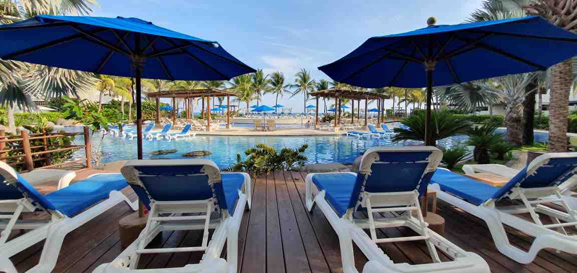 A serene pool area is presented with blue lounge chairs positioned on wooden decking. Umbrellas provide shade, while lush palm trees and greenery surround the water. The inviting pool reflects the clear sky, with glimpses of the ocean visible in the background.