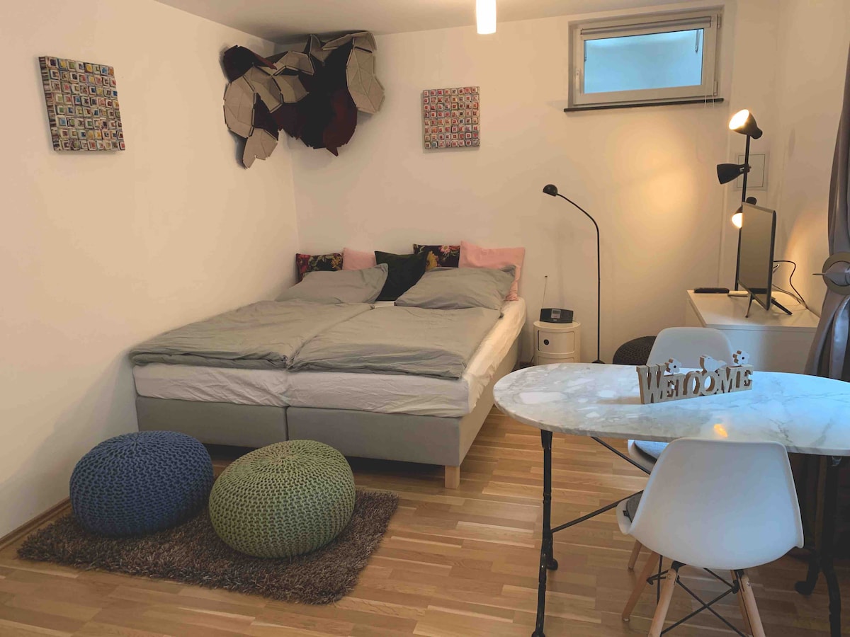 A spacious sleeping area is highlighted by a large bed with soft bedding and decorative pillows. Colorful wall art decorates the white walls. A small dining table with modern chairs sits nearby, complemented by a cozy area rug and seating poufs in vibrant colors.