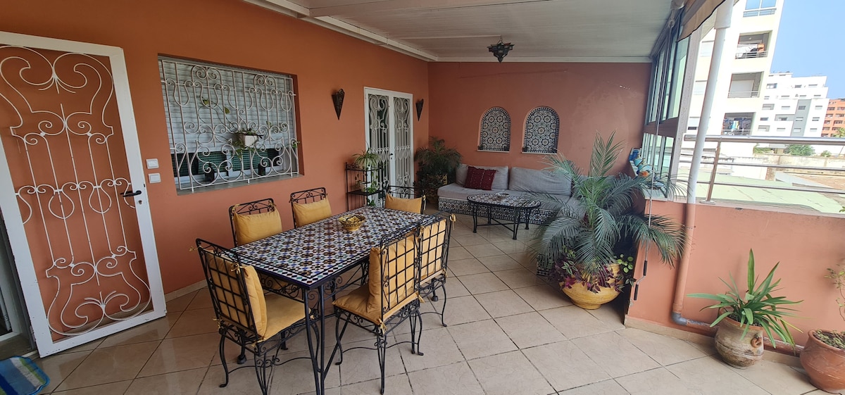 An outdoor terrace features a dining table with six metal chairs and decorative tilework. A cozy seating area consists of a sofa and lounge chairs, surrounded by potted plants. The warm orange walls add a vibrant touch, complemented by large windows allowing natural light.