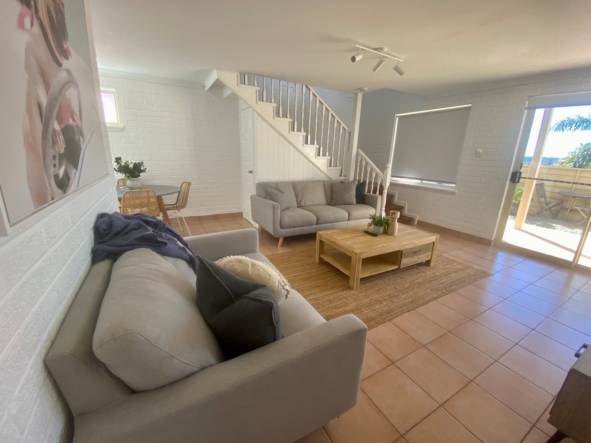 The living area features a light-filled space with two comfortable sofas positioned around a wooden coffee table. A dining table is visible in the corner, and sliding glass doors provide access to an outdoor courtyard.