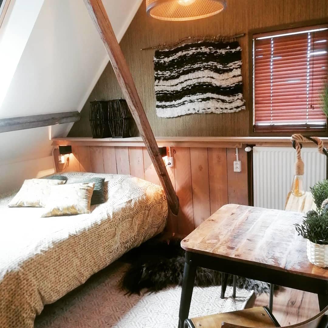 A cozy sleeping area features a textured blanket draped over a spacious bed, with decorative pillows adding detail. Warm wood tones are complemented by woven wall art. A small table sits nearby, adorned with a potted plant and natural fiber accents.