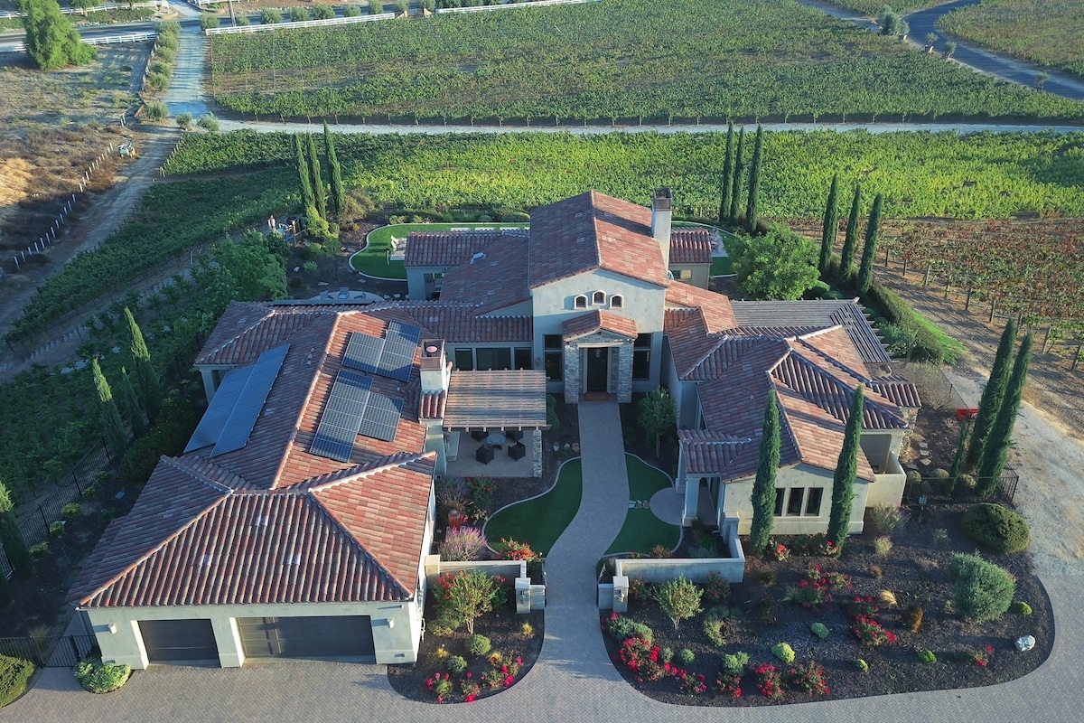 The estate is set amidst expansive vineyards, featuring a spacious layout with a combination of tile and stone pathways leading to a grand entrance. Surrounding gardens with colorful blooms and tall cypress trees add to the property's charm while showcasing its connection to the lush landscape.