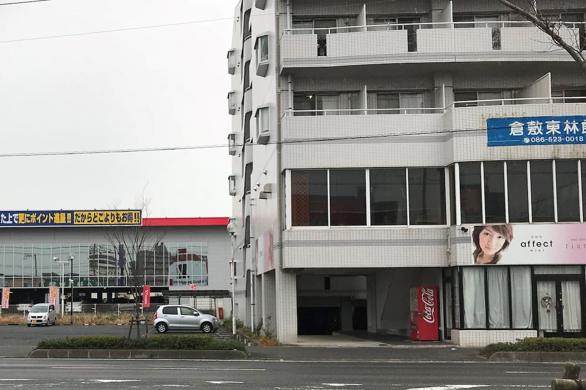 A modern building is visible, featuring multiple floors with balconies and large windows. Shops are located on the ground level, displaying signs in varying colors. A parking area is accessible, with a vehicle parked nearby. An outdoor scene captures a cloudy sky.