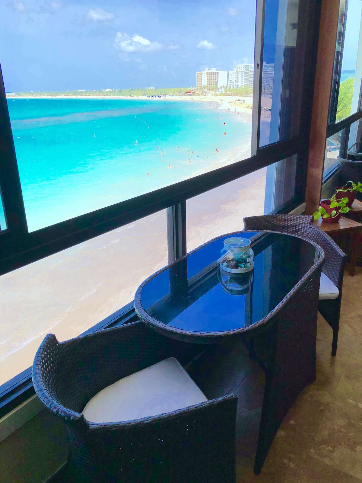A balcony area is visible, featuring two wicker chairs with light cushions and a sleek glass table. The backdrop showcases a stunning ocean view, with clear turquoise waters meeting a sandy beach. A small decorative plant is placed on the side.