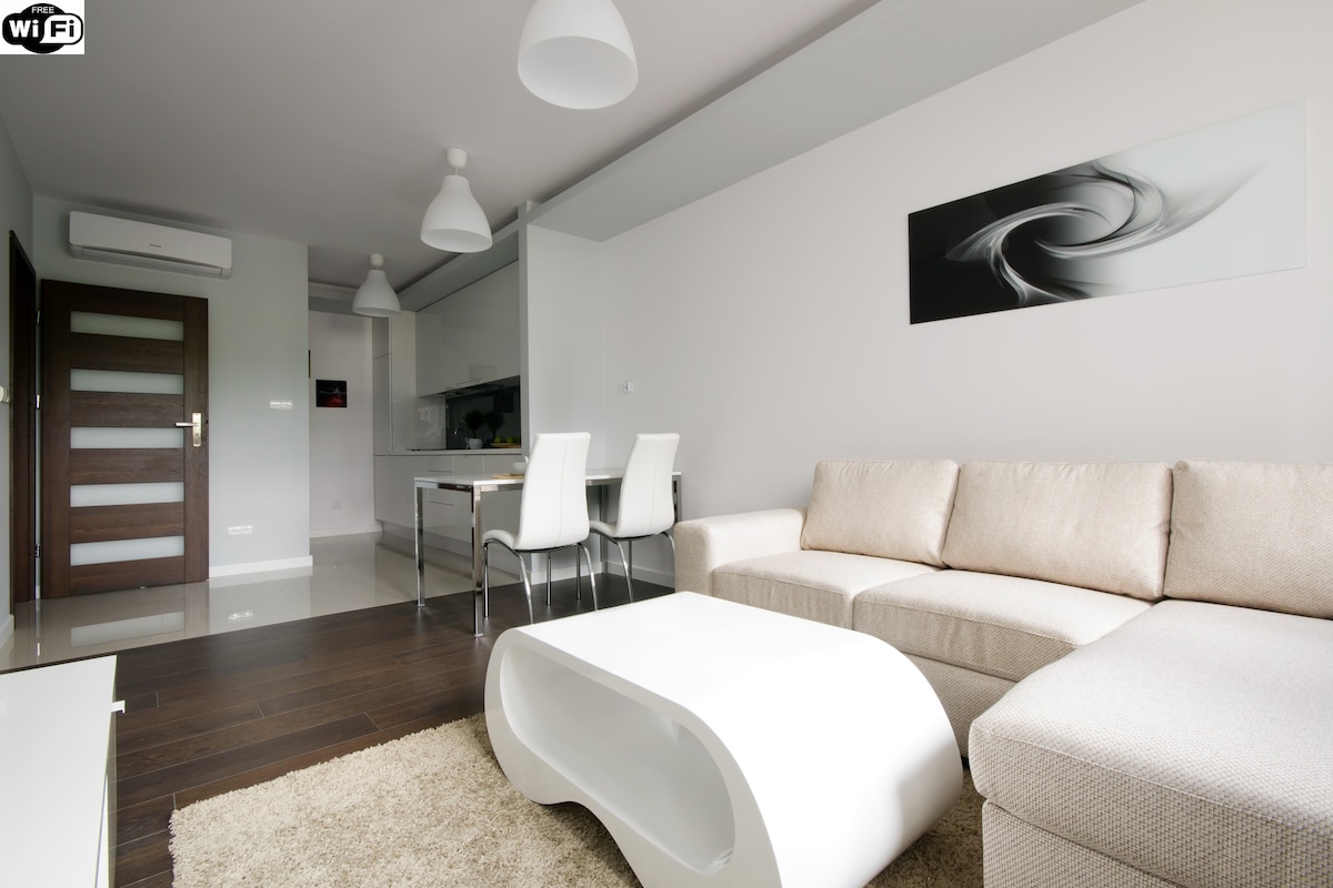 A modern living area features a light-colored sectional sofa paired with a sleek white coffee table. A dining space with four chairs is visible in the background, along with an open kitchen area. Natural light is filtered through the windows, creating a bright ambiance.