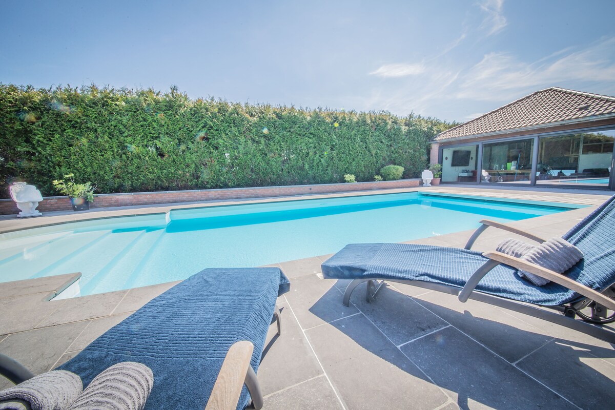 An outdoor area features a large heated swimming pool with clear blue water, surrounded by neatly trimmed hedges. Two lounge chairs in navy blue are positioned beside the pool, offering a relaxing spot to enjoy the sun.