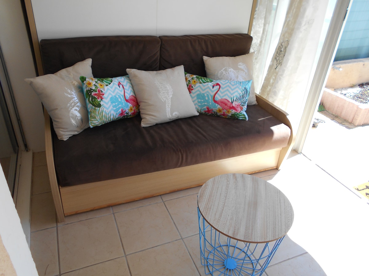 A compact seating area features a dark brown sofa adorned with colorful decorative pillows displaying tropical designs. A small round table with a wooden top and blue wire base is positioned in front. Soft light filters in through sheer curtains.
