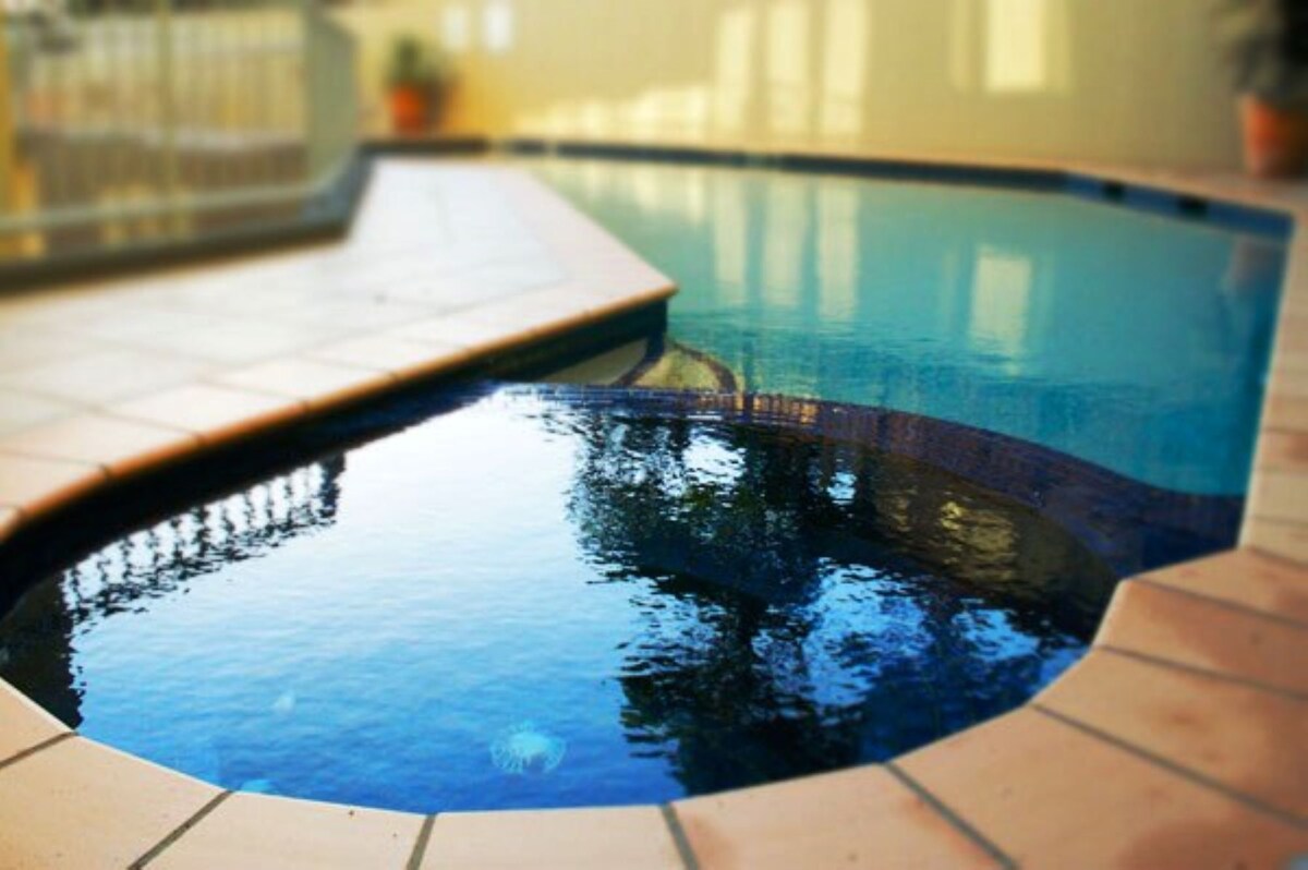 A curved pool area is showcased with terracotta tiles surrounding the water. The clear water reflects nearby foliage, creating a serene atmosphere. Sunlight glimmers on the surface, enhancing the pool's inviting appearance.