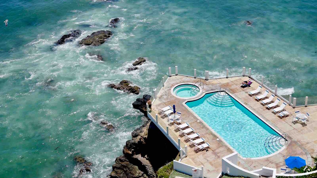 An outdoor pool area is situated on a rocky shoreline, overlooking the ocean. The pool features a separate hot tub, with multiple lounge chairs arranged nearby. Waves gently crash against the rocks, creating a serene coastal setting.