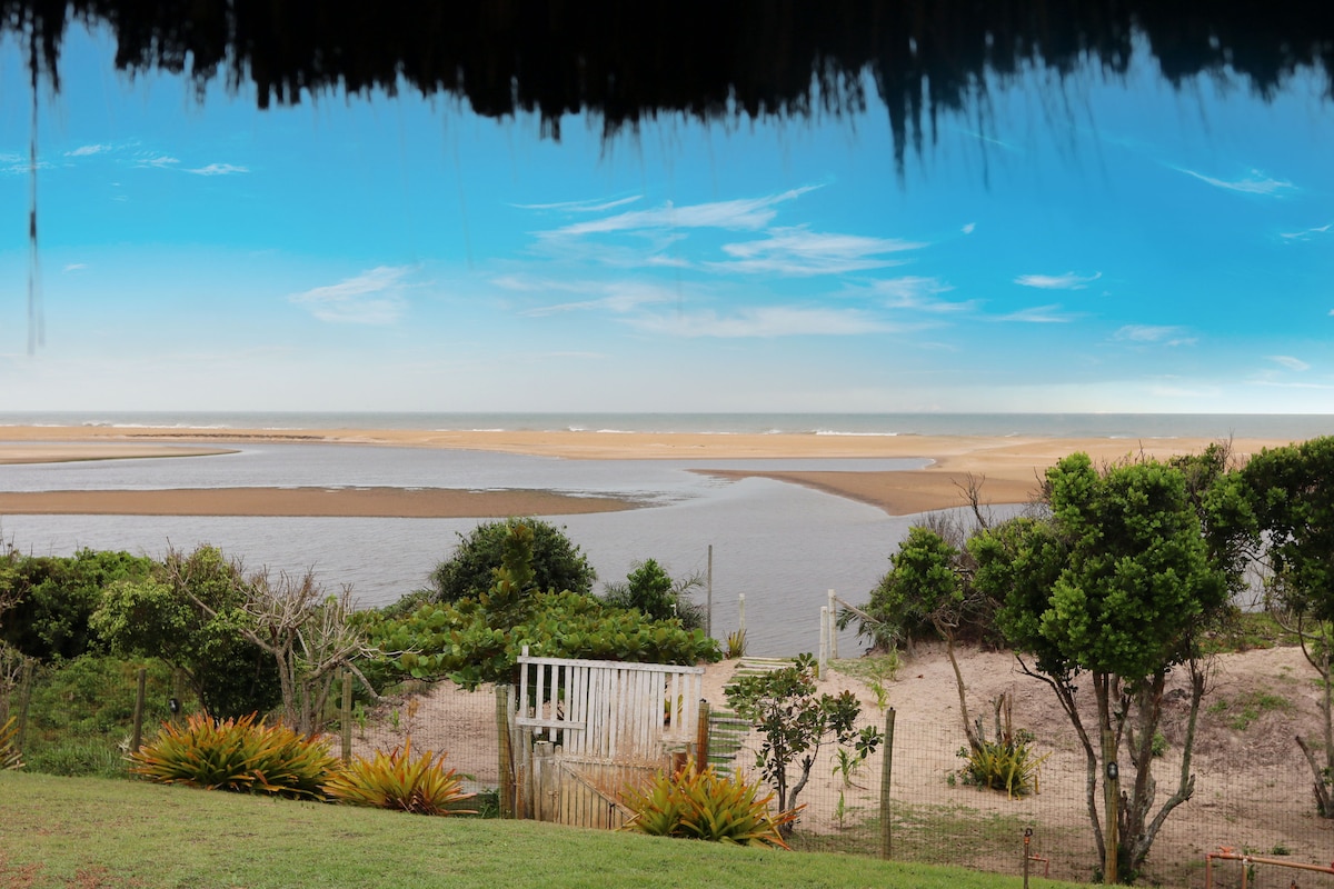 A serene view of the coastline is framed by lush greenery, showcasing a sandy beach meeting a calm river. The sky is clear with soft, blue hues, while the landscape unfolds gently, presenting a peaceful natural setting.