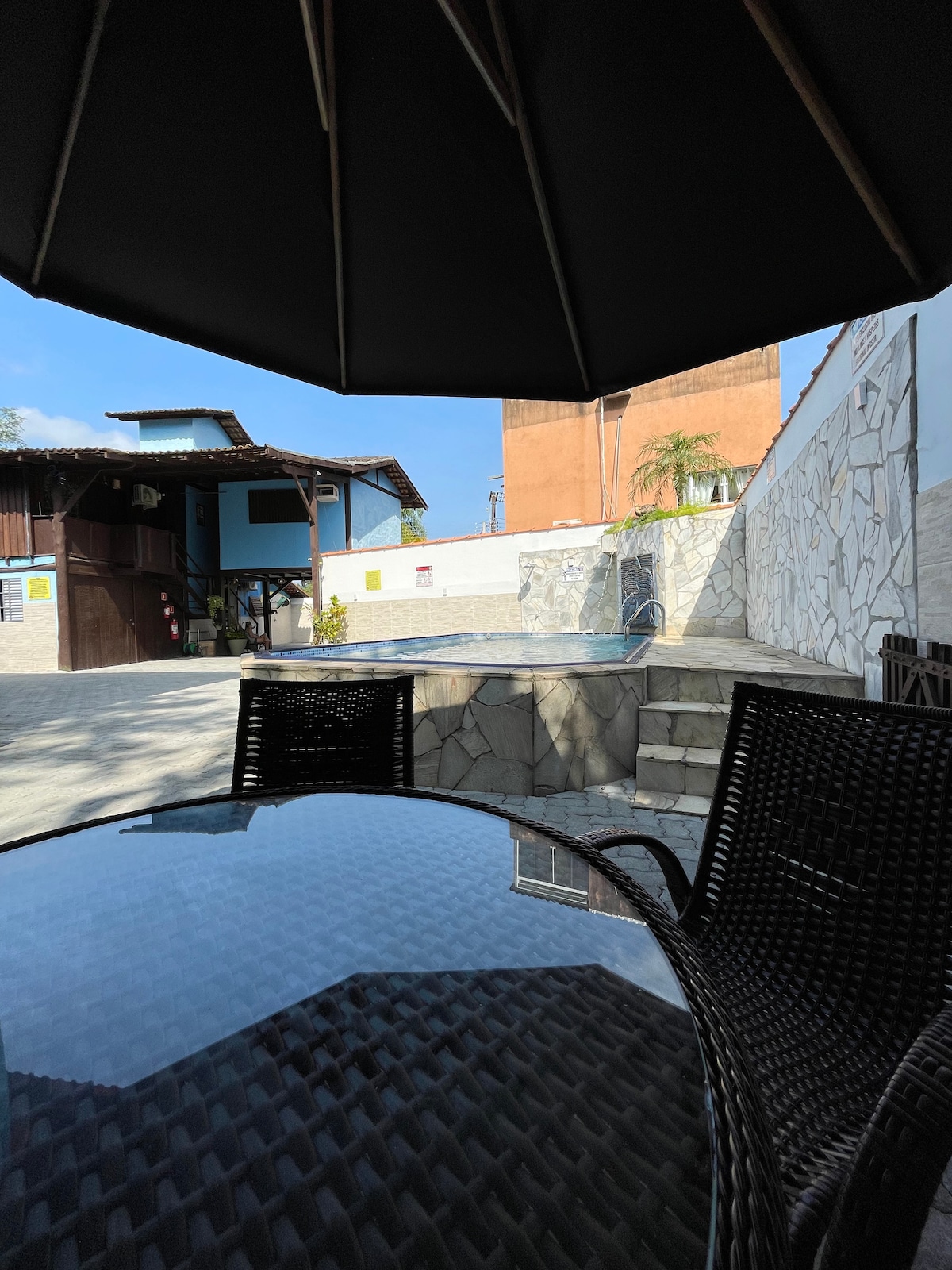 A shaded outdoor area features a glass table with wicker chairs, providing a view of the pool area. The pool is surrounded by a stone wall and visible from beneath a large umbrella that offers protection from sunlight.