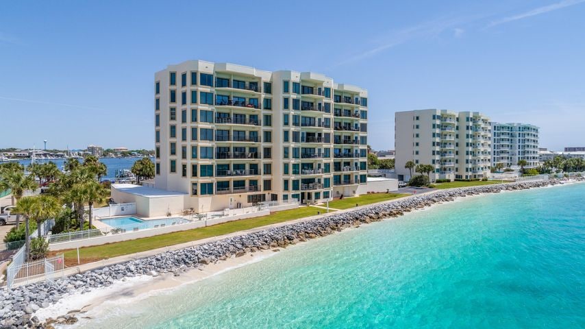 A modern condominium complex is situated along the shoreline, featuring multiple stories and ample balconies. The clear turquoise water reflects sunlight, while manicured lawns and a private pool area are visible in the foreground. The serene atmosphere is complemented by nearby palm trees.