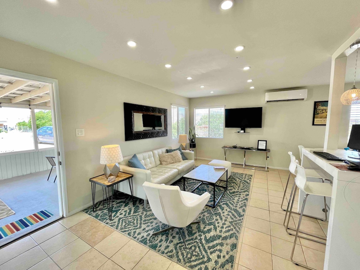 A bright living area features a light-colored sofa and modern armchairs arranged on a patterned rug. A coffee table sits at the center, while a large mirror and wall-mounted TV enhance the space. Windows allow natural light to illuminate the room.