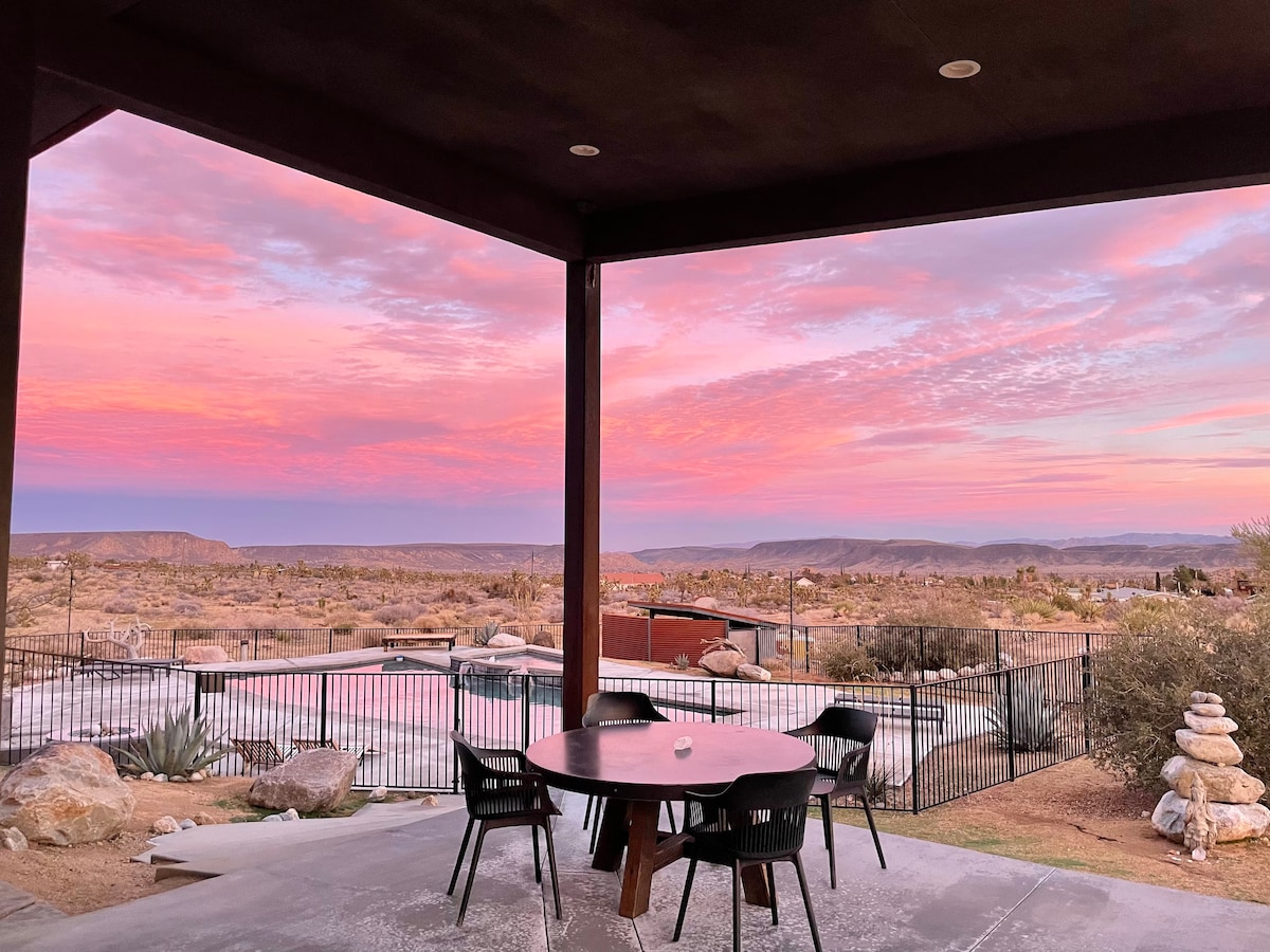 A covered outdoor dining area offers a round table surrounded by black chairs, with expansive views of desert landscapes. A soft pink and purple sky creates a serene backdrop, while the saltwater pool and natural rock formations are visible beyond the railing.