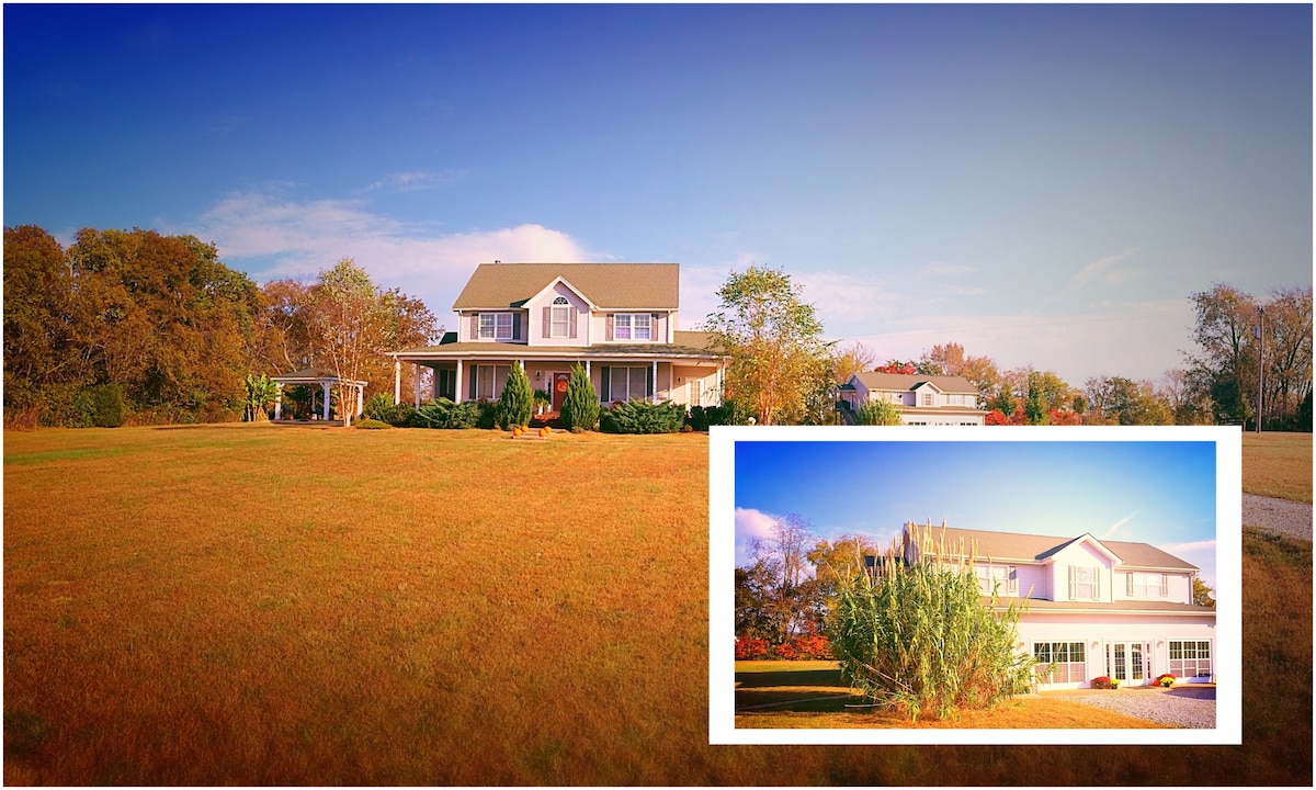 A spacious two-story house is situated on a large grassy expanse, surrounded by trees. The house features a welcoming porch and multiple windows that reflect natural light. An inset image showcases a closer view, highlighting the inviting exterior and landscaped areas.