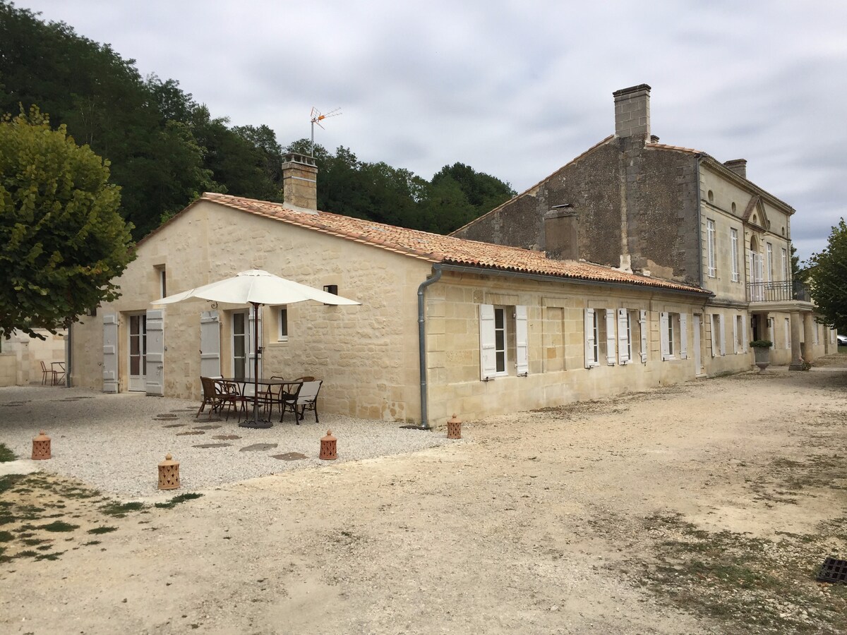 A charming stone building is set against a backdrop of gently rolling hills. A spacious terrace with outdoor dining furniture is visible, accompanied by an umbrella for shade. The gravel pathway leads towards the entrance, bordered by greenery.