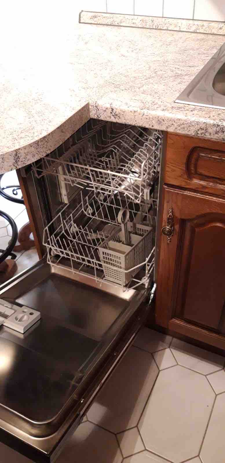 A spacious kitchen corner is shown with an open dishwasher, featuring two racks designed for dishes and utensils. The granite countertop complements the wooden cabinetry, and tiled flooring is visible underneath. The nearby sink is partially highlighted, integrating seamlessly into the kitchen layout.