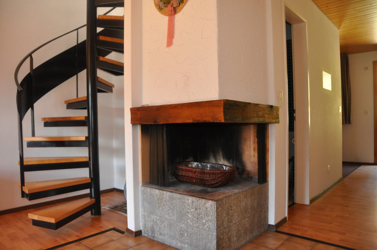 A spiral staircase made of wood and metal leads to the upper level. A stone fireplace with a wooden mantel is placed on the floor, offering a central focal point to the room. Natural light from a nearby window softly illuminates the space.