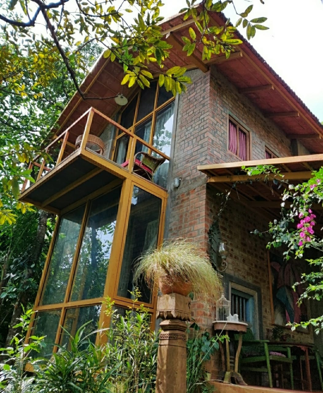 A charming brick house is framed by lush greenery, featuring large glass windows and a spacious balcony adorned with potted plants. The roof has a slight overhang, and flora enhances the natural ambiance surrounding the home.