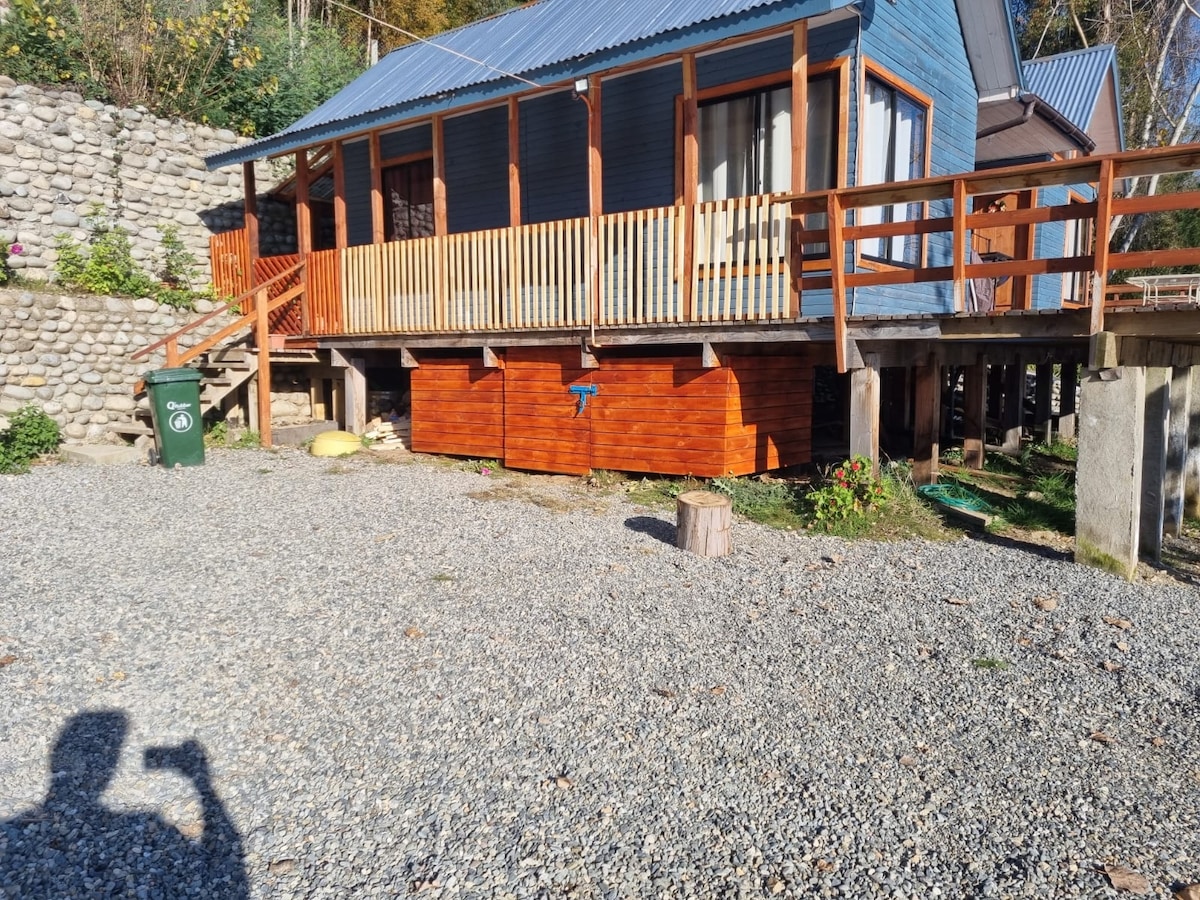 A charming cabin is set on elevated stilts, featuring a wooden deck with a railing that provides access to an outdoor space. The gravel area below is neatly arranged, with green plants and trees in the background, adding to the natural setting.