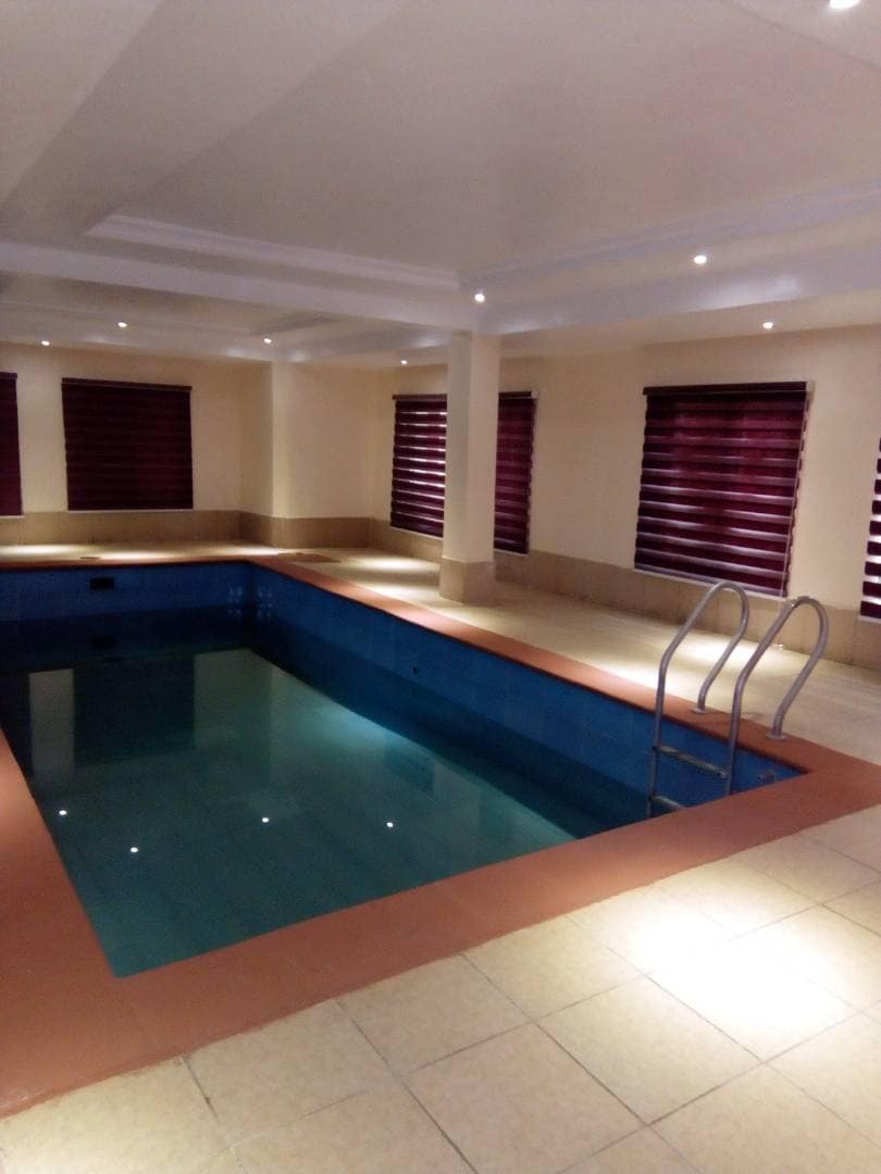 An indoor swimming pool is framed by tiled flooring and features a gentle slope leading to steps for easy entry. Large windows with dark blinds allow for natural light, while recessed lighting adds a warm glow to the space.